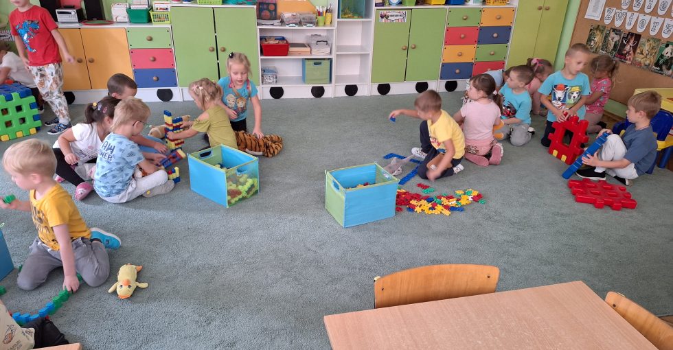 Dzieci siedzą na dywanie i budują z różnych klocków domy dla zwierząt.