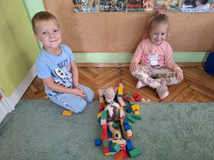 Dziewczynka i chłopiec siedzą na dywanie i prezentują swoją budowlę z klocków - dom dla zwierząt egzotycznych.