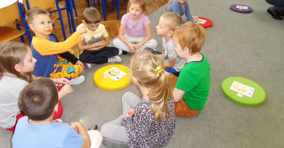 grupa dzieci siedzi wokół emotki z uśmiechem i kolejno opowiadają dlaczego wybrały taki symbool.