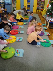Dzieci siedzą na dywanie i układają cyfry na kołach-poduszkach tworzących tarcze zegarowe.