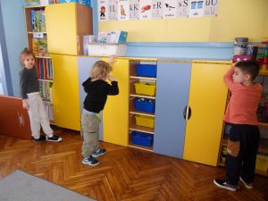 Troje dzieci mierzy taśmą mierniczą długość ustawionych pod ścianą szafek.