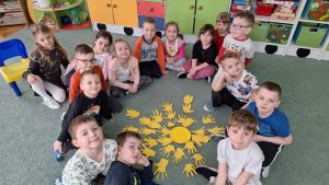 Dzieci z grupy czwartek pozują do wspólnej fotografii z wykonaną przez siebie pracą plastyczną - wyciętymi dłońmi z żółtego papieru, ułożonymi wokół żółtego talerzyka. Kompozycja tworzy słońce. 