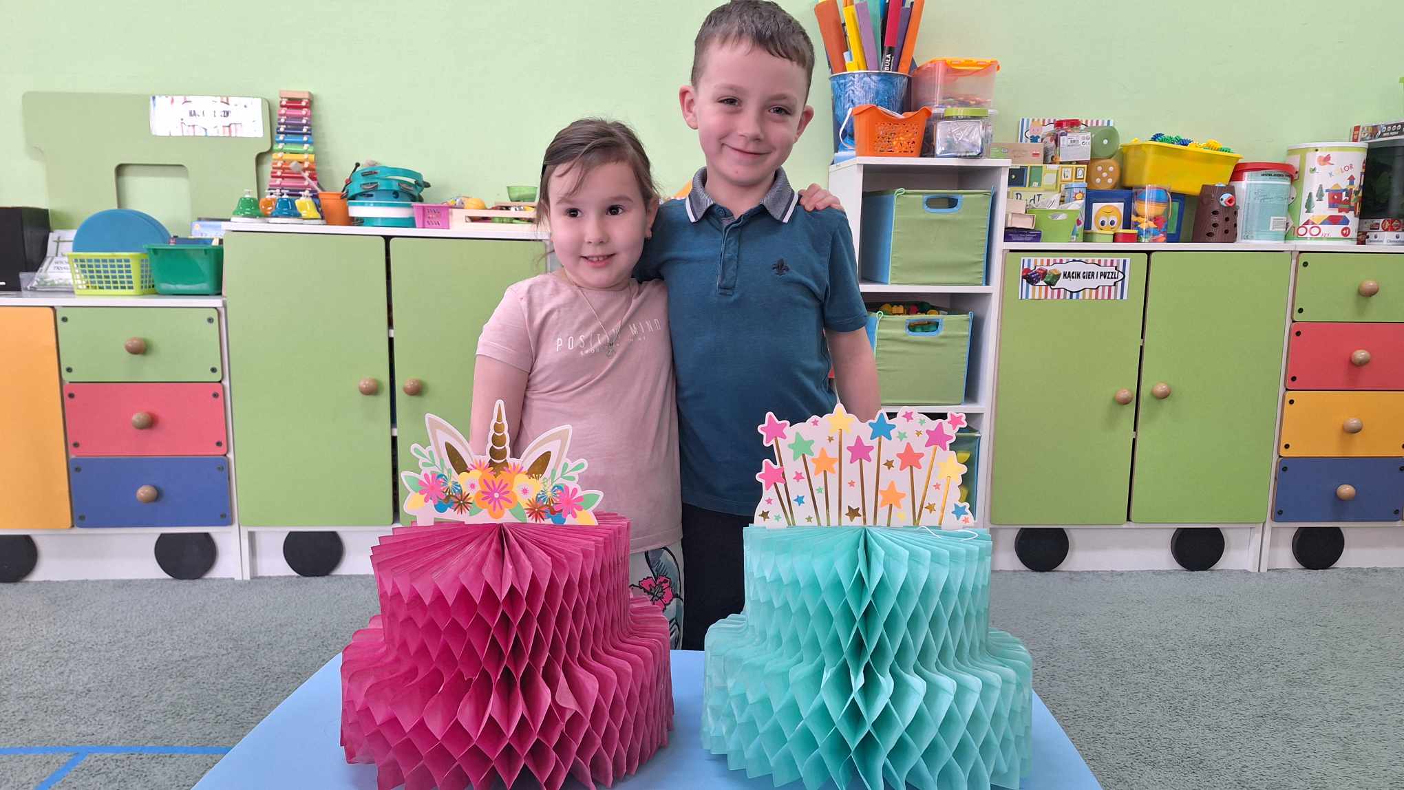 Antosia i Oskar pozują do zdjęcia obejmując się. Przed nimi stoi stolik z dwoma papierowymi tortami - różowym i niebieskim.