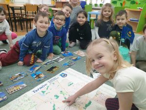 Dzieci siedzą wokół mapy Tarnowa, a dziewczynka wskazuje miasto na mapie.