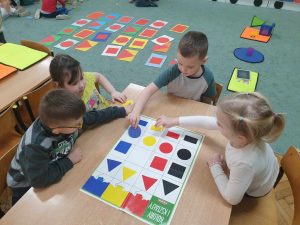 Czworo dzieci siedzi przy stoliku i uzupełnia planszę odpowiednimi figurami geometrycznymi, zwracając uwagę na odpowiedni kształt i kolor.