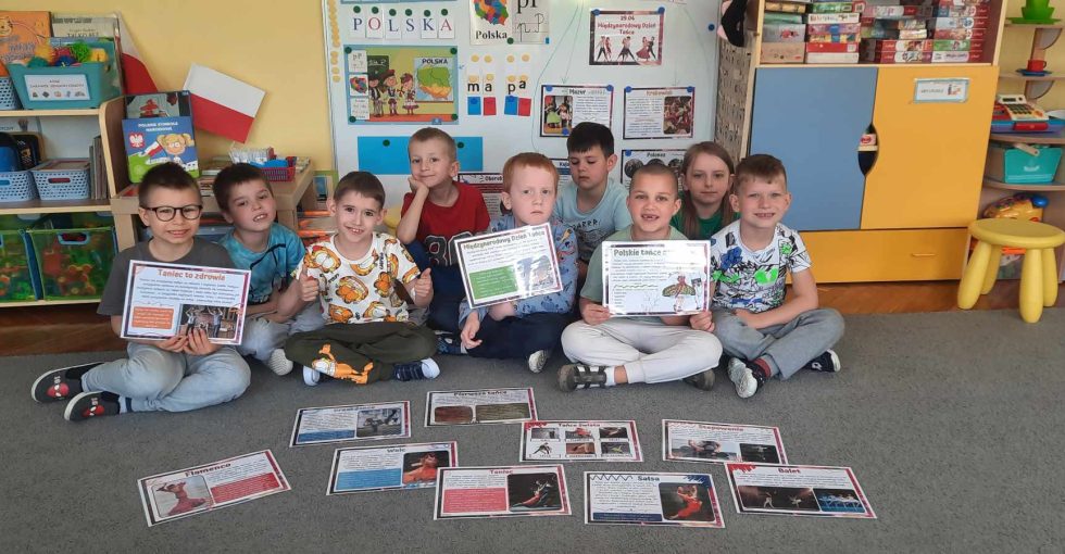 Dzieci siedzą na dywanie, pozują do zdjęcia trzymając ilustracje i grafiki opisujące style tańca z całego świata.