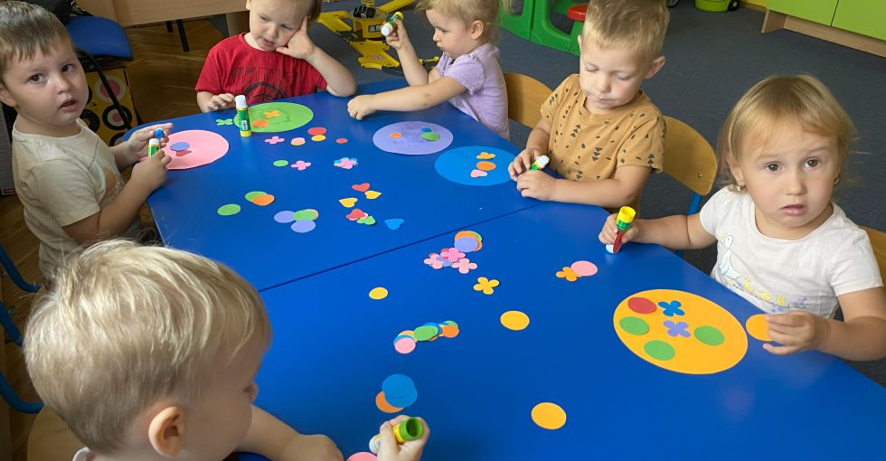 Dzieci siedzą przy stolikach na których leżą powycinane z kolorowego papieru ozdoby (kwiatuszki, kółka,serduszka)