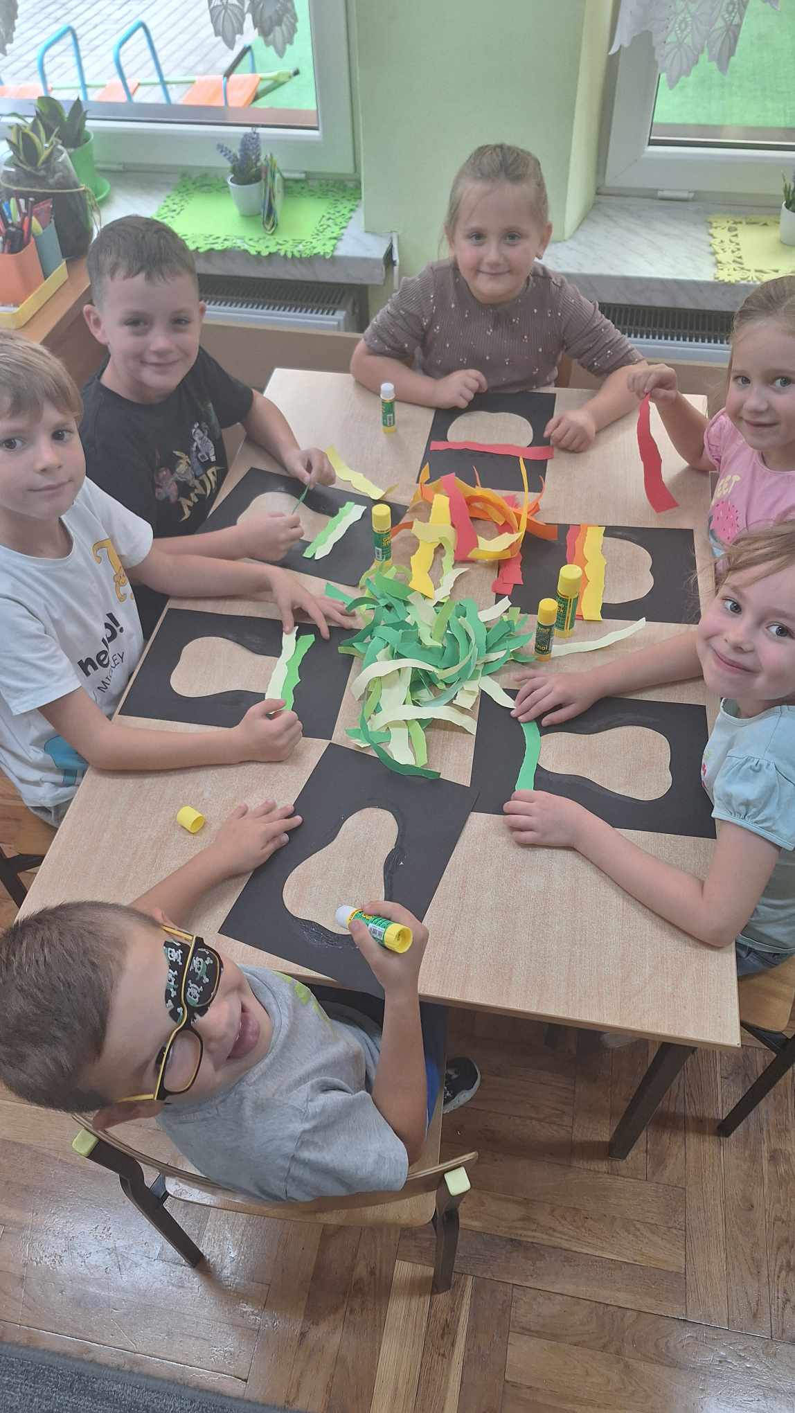 Dzieci siedzą przy stole, przed każdym leży czarny szablon z jabłkiem lub gruszką. Na środku stoją kolorowe paski papieru oraz kleje.