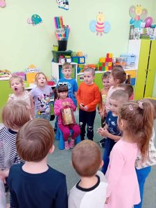 Solenizantka siedzi na krzesełku. W rękach trzyma torbę z cukierkami. Wokół stoją dzieci i śpiewają "Sto lat".