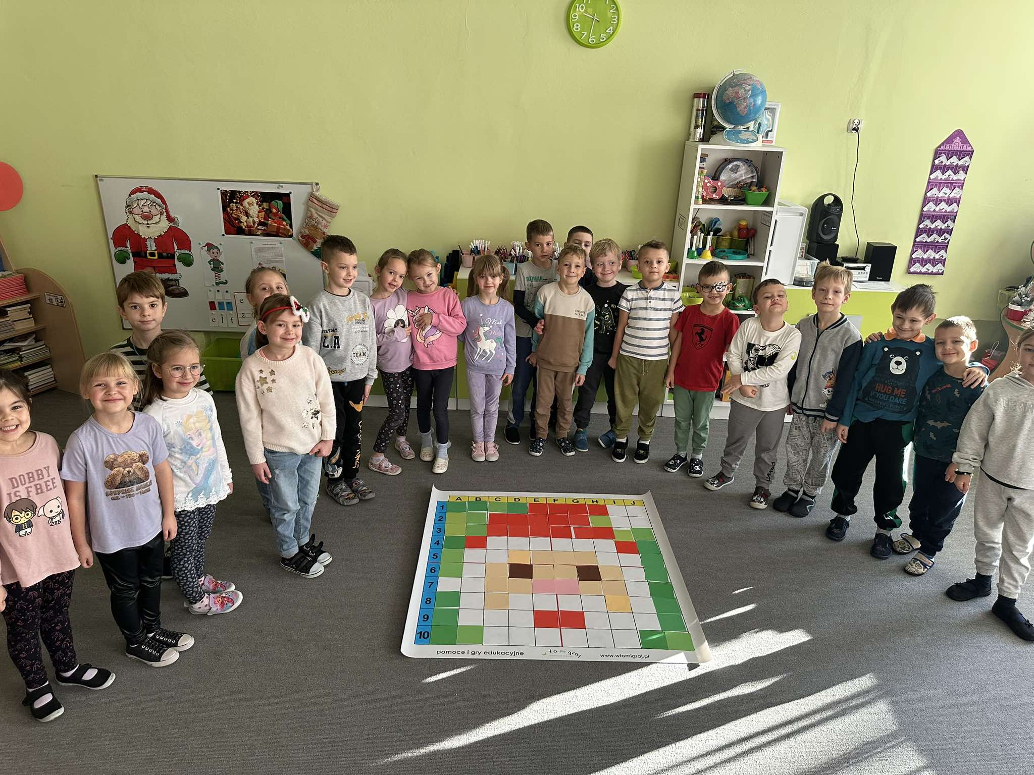 Dzieci z grupy V stoją wokół maty do kodowania, na której ułożyli twarz świętego mikołaja, używając kolorowych katrtoników.