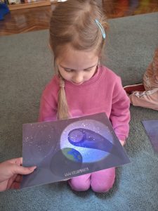 Dziewczynka siedzi na dywanie. W rękach trzyma ilustrację ciemnego nieba-kosmosu. Podświetlają ilustrację latarką by zobaczyć ukryty elementy kosmosu - meteoryt i słońce.