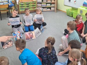 Troje dzieci trzyma w rękach ilustracje z zimowymi pejzażami. Przed nimi na małych sztalugach stoją ilustracje przedstawiające: martwą naturę, abstrakcję, portret i pejzaż.