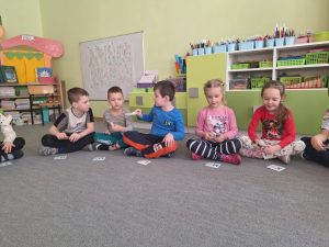Grupa dzieci siedzi na dywanie. Za pomocą spinaczy przekazują sobie kartę z literą.
