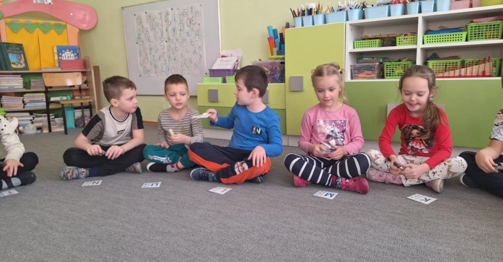 Grupa dzieci siedzi na dywanie. Za pomocą spinaczy przekazują sobie kartę z literą.