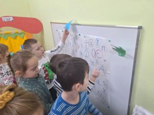 Grupa dzieci stoi przed suchościeralną tablicą. Przy pomocy magnesów przyczepiają do niej swoje prace.