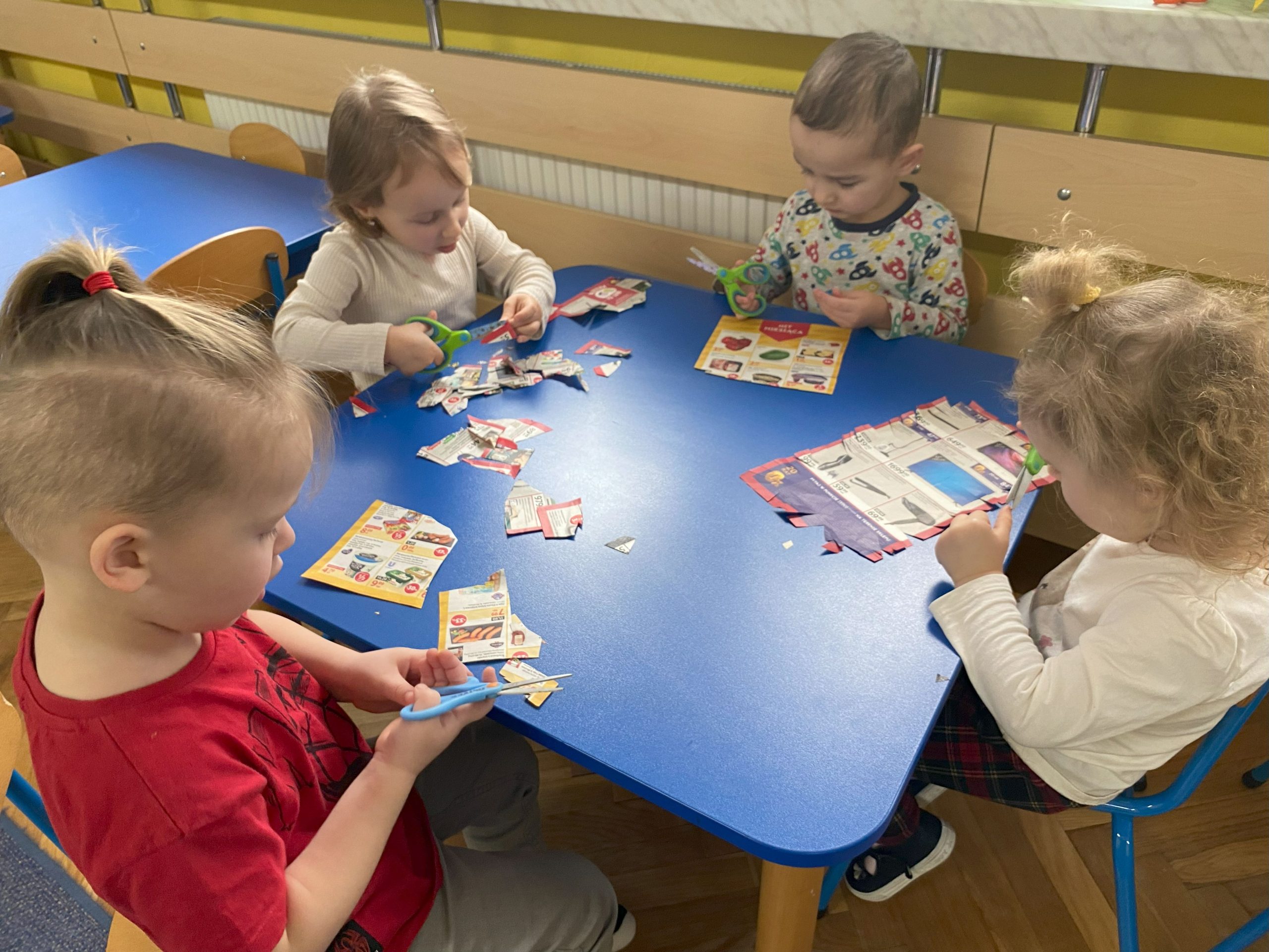 czwórka przedszkolaków tnie nożyczkami gazetki reklamowe