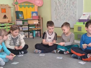 Grupa dzieci siedzi na dywanie. Za pomocą spinaczy przekazują sobie kartę z literą.