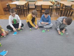Grupa dzieci siedzi na dywanie. Mają przed sobą dłoń wycięta z papieru. Przy użyciu kleju przyklejają do poszczególnych palców wybrane przez siebie samogłoski.