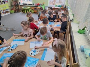 Grupa dzieci siedzi przy stolikach 6 osobowych. Każdy wykonuje pracę plastyczną - przy użyciu  różnych odcieni niebieskich farb maluje zimowy pejzaż.