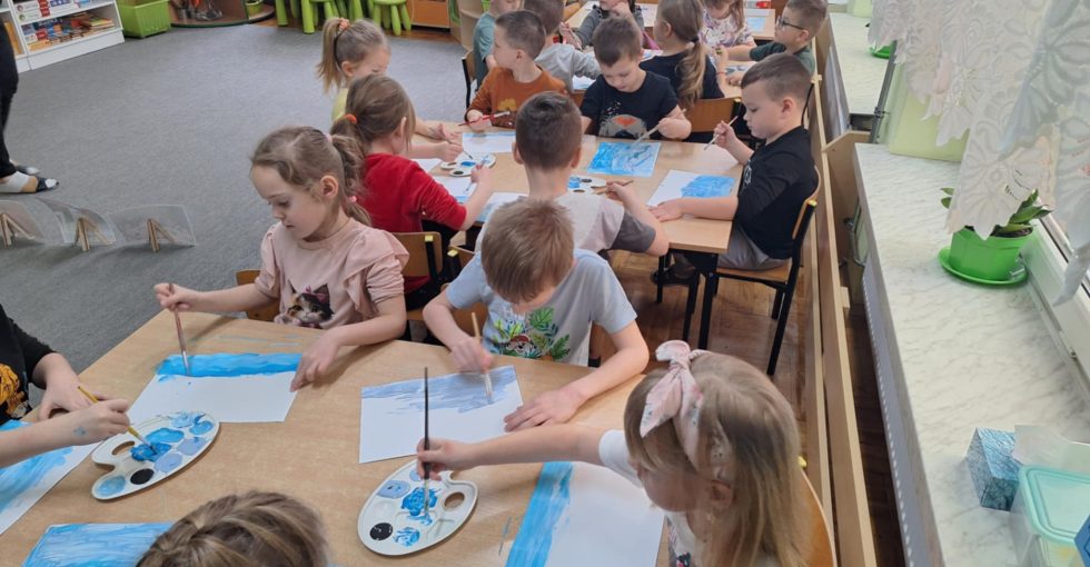 Grupa dzieci siedzi przy stolikach 6 osobowych. Każdy wykonuje pracę plastyczną - przy użyciu różnych odcieni niebieskich farb maluje zimowy pejzaż.