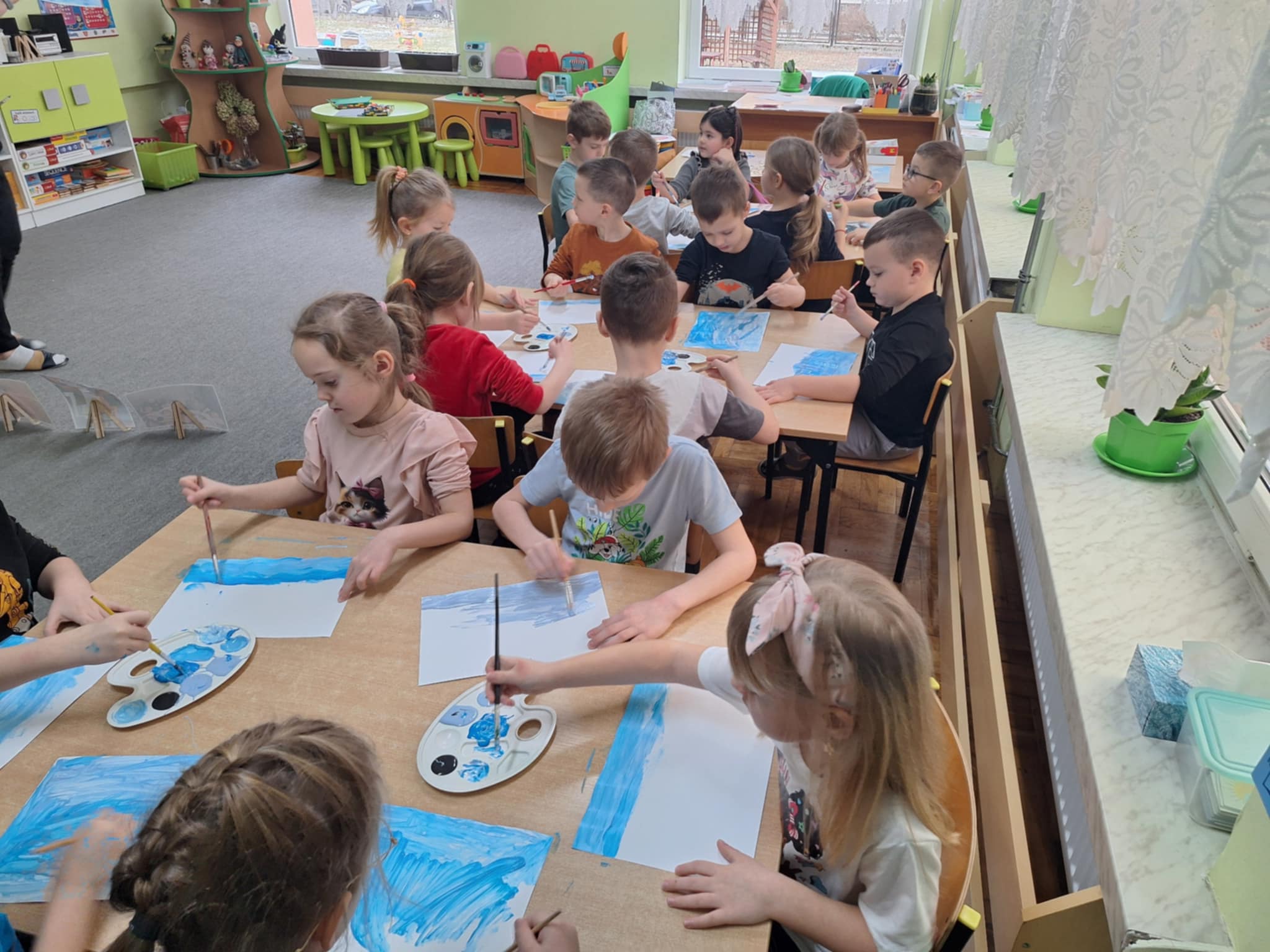 Grupa dzieci siedzi przy stolikach 6 osobowych. Każdy wykonuje pracę plastyczną - przy użyciu różnych odcieni niebieskich farb maluje zimowy pejzaż.