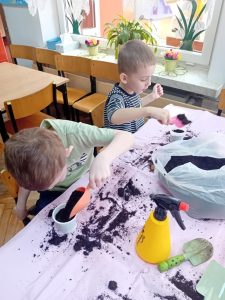 Dwóch chłopców siedzi przy stoliku. Wsypują za pomocą łopatek ziemię do plastikowych pojemników.