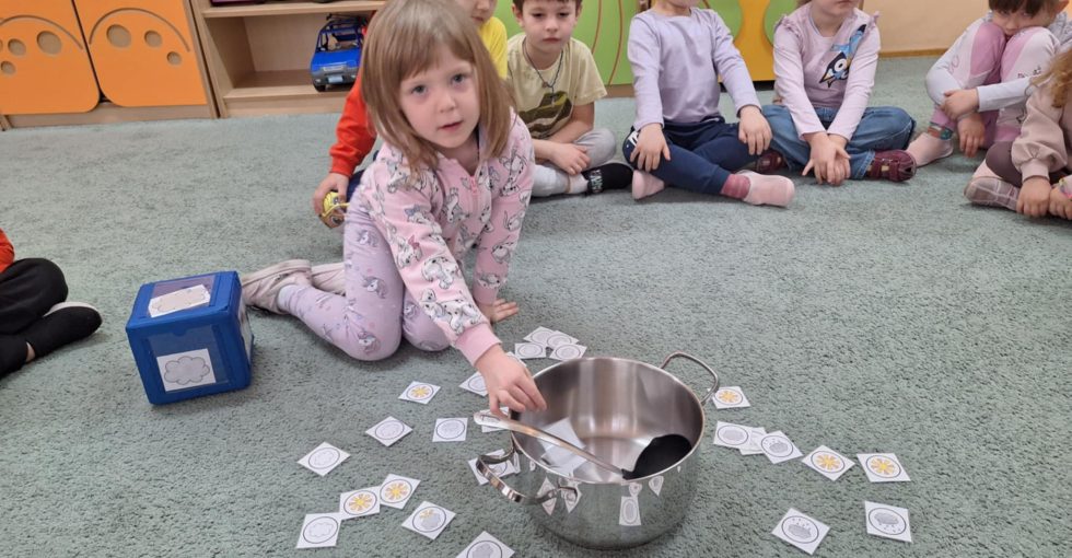 Dziewczynka miesza chochlą w garnku. Wokół leżą emblematy pogodowe. W tle siedzą dzieci z grupy.