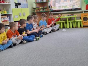 Grupa chłopców siedzi w rzędzie. Trzyma w rękach sznurek, po którym nawlekają duże korale.