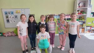Solenizant siedzi na krześle. w rękach trzyma papierowy tort. za nim stoją koleżanki z grupy.