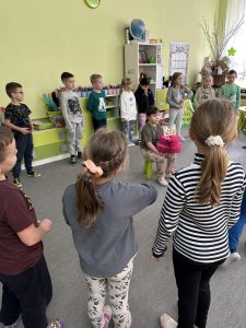 Solenizatka siedzi w kole na krzesełku. Trzyma papierowy tort. Wokół stoją dzieci z grupy.