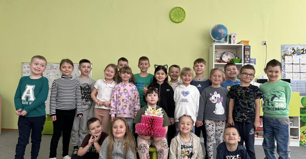Solenizantka siedzi na środku. Na kolanach trzyma papierowy tort. Dzieci z grupy są ustawione za nią i przed nią. Wszyscy pozują do zdjęcia.