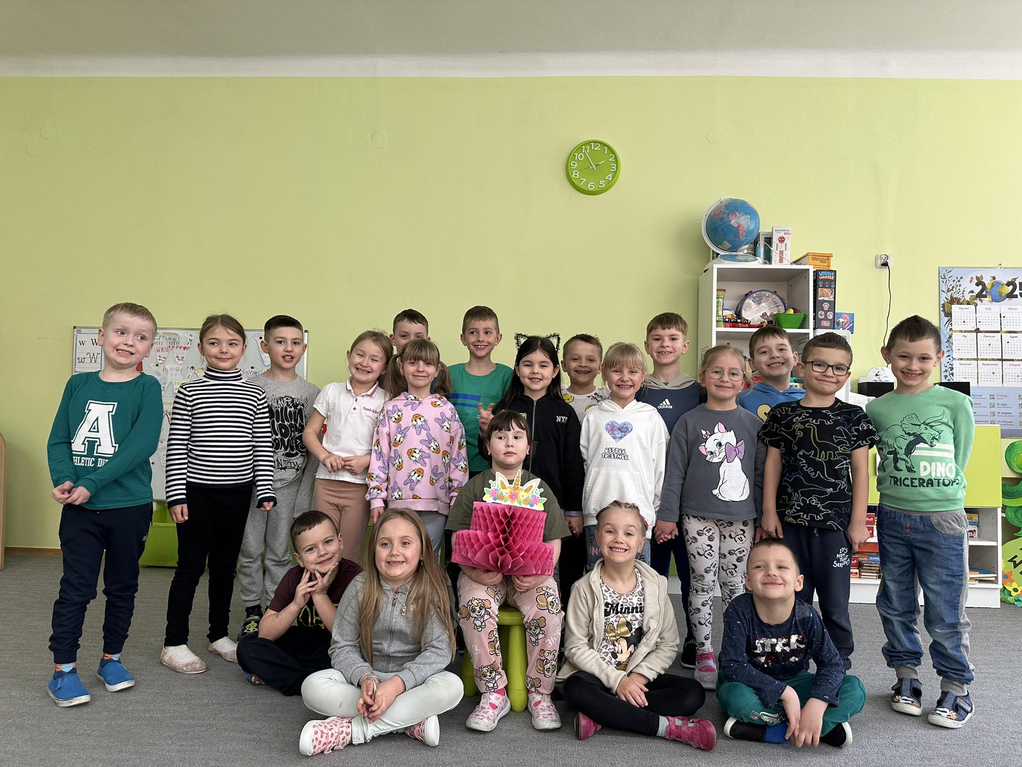 Solenizantka siedzi na środku. Na kolanach trzyma papierowy tort. Dzieci z grupy są ustawione za nią i przed nią. Wszyscy pozują do zdjęcia.