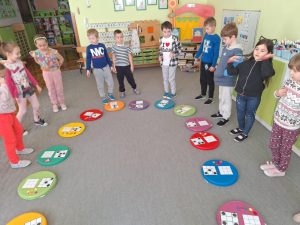 Dzieci stoją w kole. Przed nimi leżą kolorowe podkładki na których leżą przygotowane zadania do wykonania.