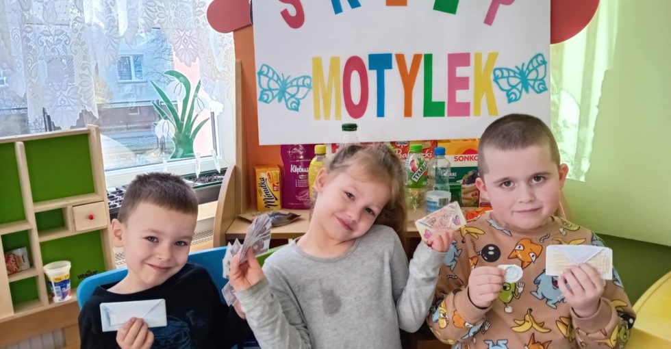 Trójka dzieci siedzi na krześle przed szyldem przedszkolnego sklepiku. Dzieci prezentują wykonane własnoręcznie papierowe portfele i pieniążki.