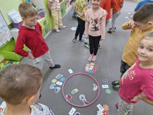 Dzieci stoją w grupach 4osobowych. W środku  rozłożone są emblematy wiosenne i kartki z cyframi.