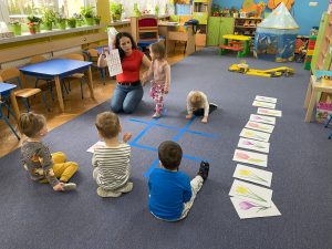 nauczycielka pokazuje dziewczynce schemat ułożenia kwiatków, na dywanie przyklejona jest taśma tworząca trzy pola poziome i trzy pionowe