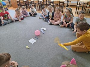 Dzieci siedzą w kole. W środku leżą przedmioty. Jeden chłopiec próbuje wprawić je w ruch przy użyciu wachlarza.