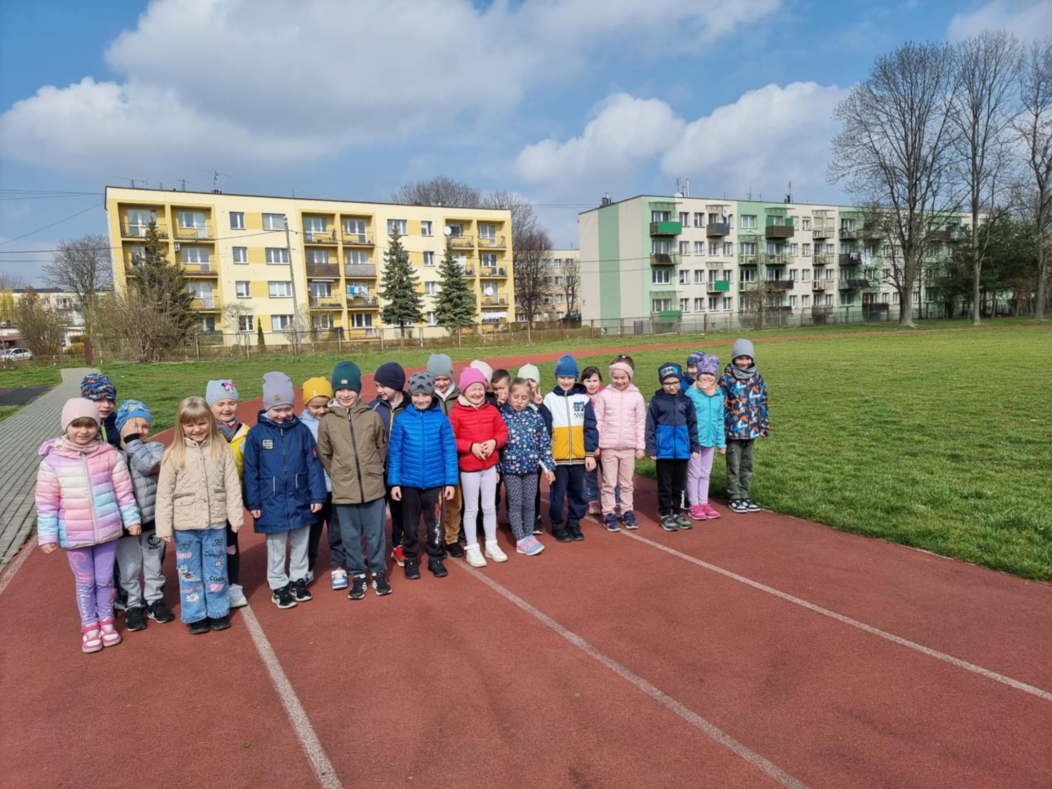 Grupa dzieci stoi na bieżni wokół boiska szkolnego.