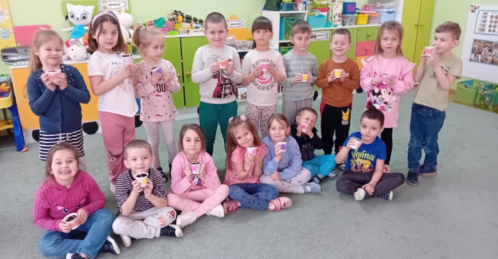 Część dzieci siedzi na dywanie a część stoi za nimi. Dzieci trzymają w rękach swoje posadzone cebulki w ozdobionych kolorowymi naklejkami pojemnikach.