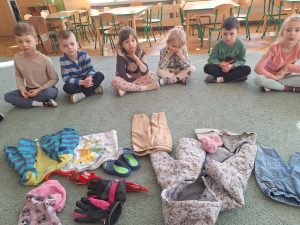 Dzieci siedzą w kole na dywanie. W środku leżą różnorodne ubrania.