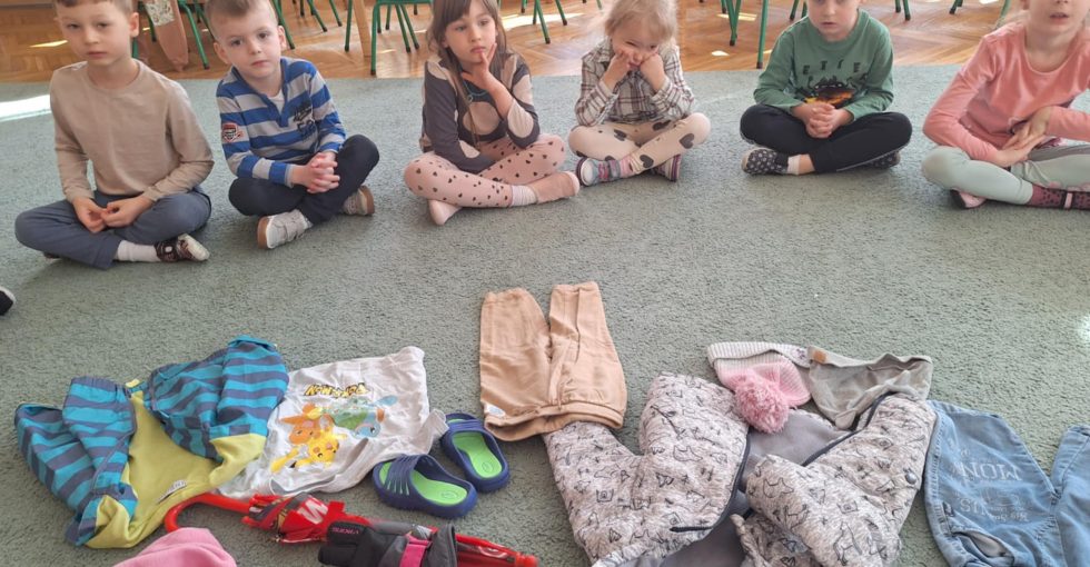 Dzieci siedzą w kole na dywanie. W środku leżą różnorodne ubrania.