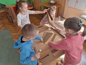 Grupa dzieci przykleja brązowe paski do kartonu w kształcie domku.