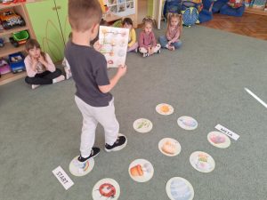 Na dywanie rozłożone są wiosenne emblematy. Dziecko porusza się po nich zgodnie z obrazkową instrukcją trzymaną w rękach.