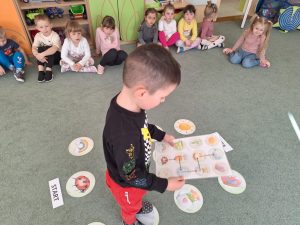Na dywanie rozłożone są wiosenne emblematy. Dziecko porusza się po nich zgodnie z obrazkową instrukcją trzymaną w rękach.