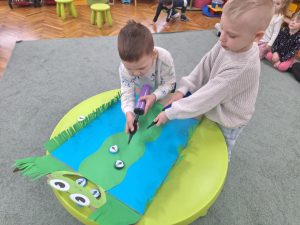 Dwójka dzieci trzyma pompki do balonów. Przesuwają muchy zrobione z zakrętek, po kolorowej trasie. Karmią żabkę.