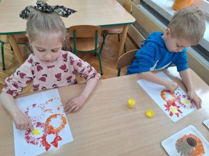 Dwoje dzieci przy stoliku. Wykonują prace plasytyczną - odbijają nacięte rolki po papierze tworząc wiosenne kwiaty.