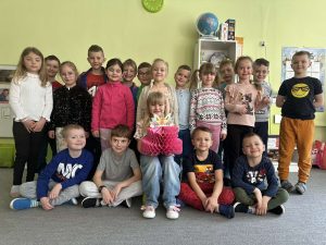 Solenizantka siedzi na krzesełku. Trzyma w rękach papierowy tort. Wokół niej stoją i siedzą pozostałe osoby z grupy.
