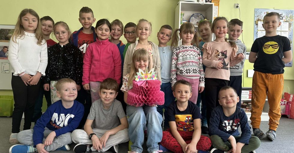 Solenizantka siedzi na krzesełku. Trzyma w rękach papierowy tort. Wokół niej stoją i siedzą pozostałe osoby z grupy.