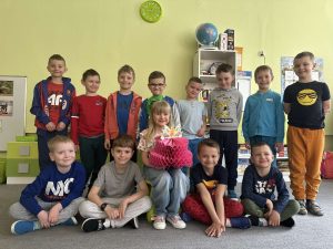Solenizantka siedzi na krzesełku. Trzyma w rękach papierowy tort. Wokół niej stoją i siedzą pozostałe chłopcy z grupy.