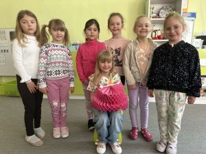 Solenizantka siedzi na krzesełku. Trzyma w rękach papierowy tort. Wokół niej stoją pozostałe dziewczynki z grupy.